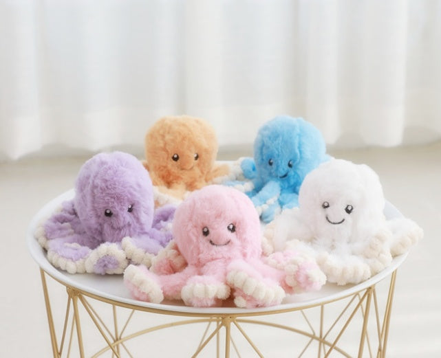 Five colorful plush octopus toys on a small round table with a white curtain background.
