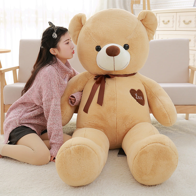 Large beige teddy bear with a heart and bow tie held by a person in a cozy living room.