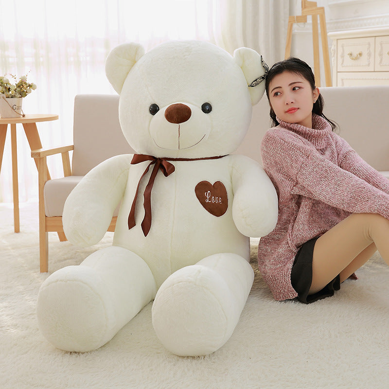 Large white teddy bear with a brown bow and heart on a couch next to a person wearing a pink sweater.