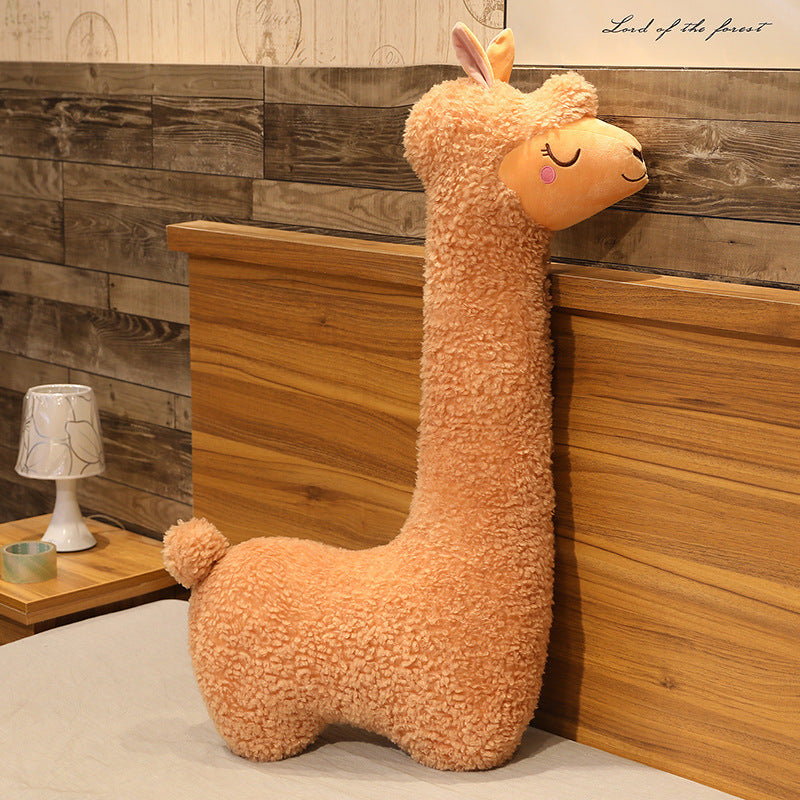 Plush llama toy leaning against a wooden wall with a lamp and small table in the background.
