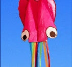 Pink kite with a face and colorful tail against a blue sky.