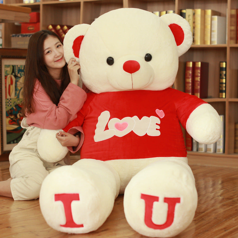 Person holding a large white teddy bear with a red shirt that says I LOVE U in a room with bookshelves.