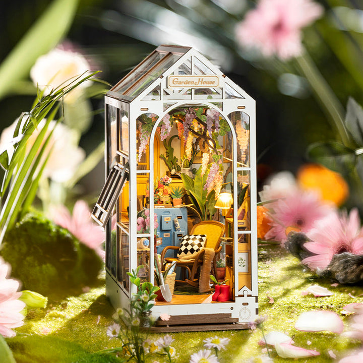 Miniature model of a garden house with indoor plants and furniture, surrounded by flowers and greenery.