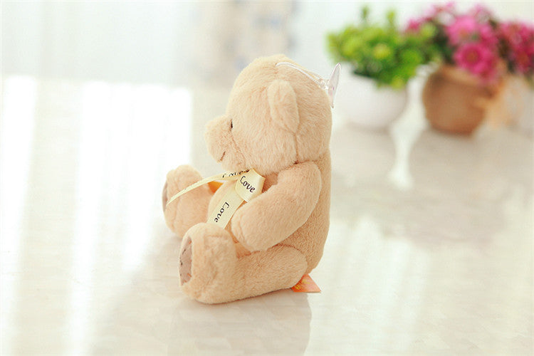 Plush teddy bear with a heart-shaped bow on a white surface with flowers in the background.