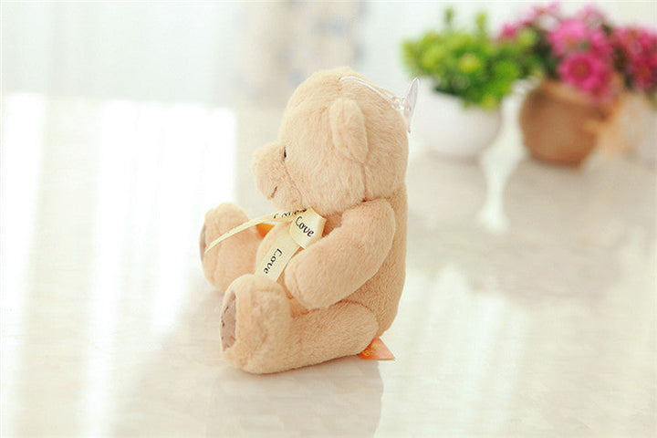 Plush teddy bear with a heart-shaped bow on a white surface with flowers in the background.