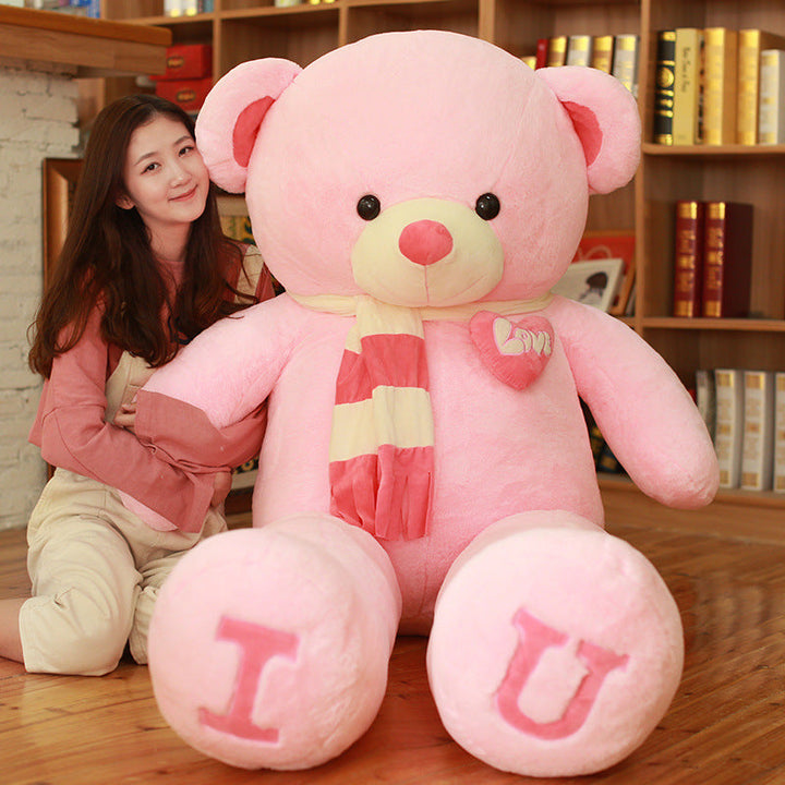 Large pink teddy bear with I U on paws, sitting next to a person in a room with bookshelves.