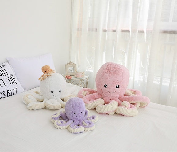 Three plush octopus toys in white, pink, and purple on a bed with a light background.