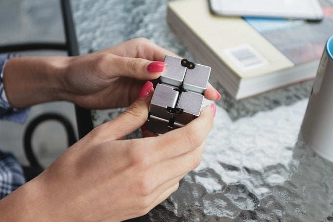 Person holding a metallic fidget cube on a blurred outdoor background.