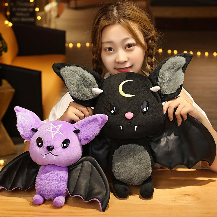 Person holding two bat-shaped plush toys, one purple and one black with a moon design, in a warm indoor setting.