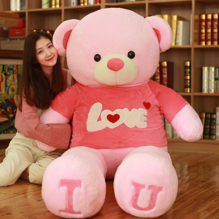 Large pink teddy bear with I U on a wooden floor in front of a bookshelf.