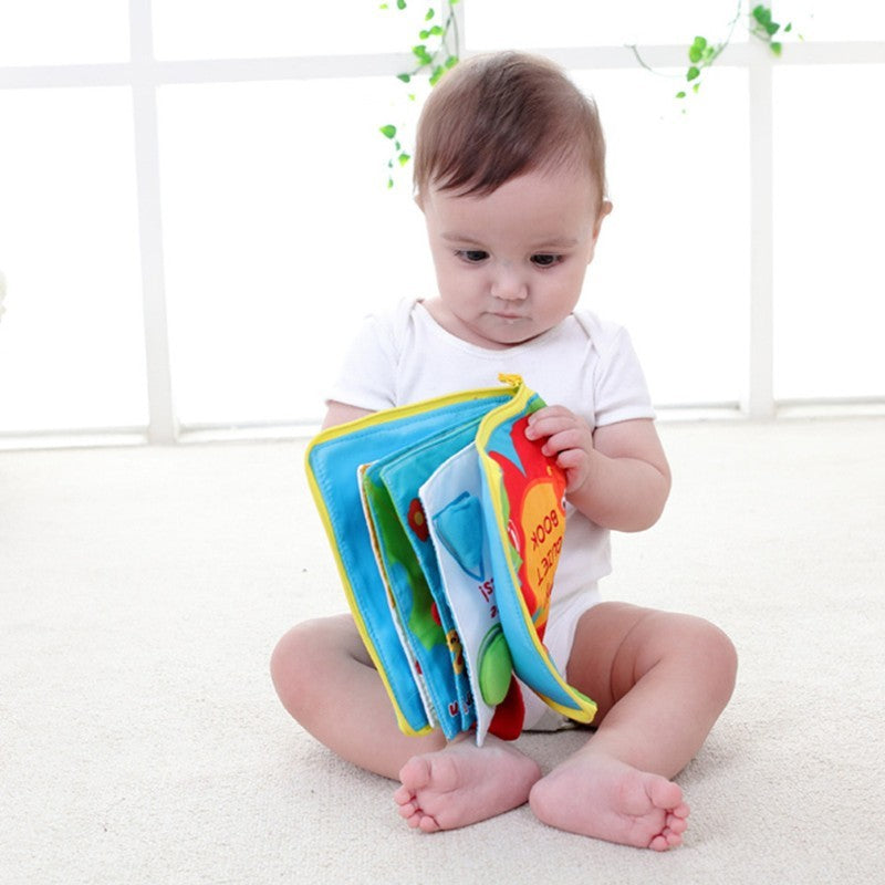 Baby holding a colorful book on a white floor with a white wall and green plants in the background.