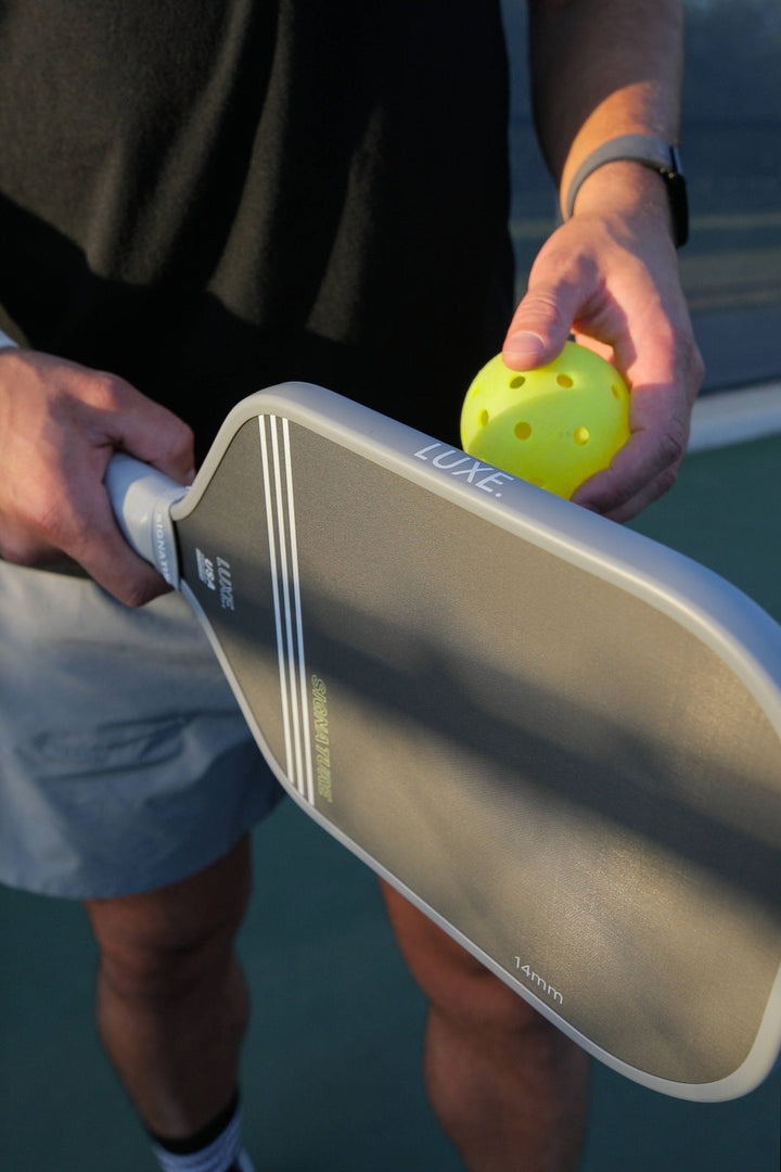 Person holding a pickleball paddle and a yellow pickleball on a court.