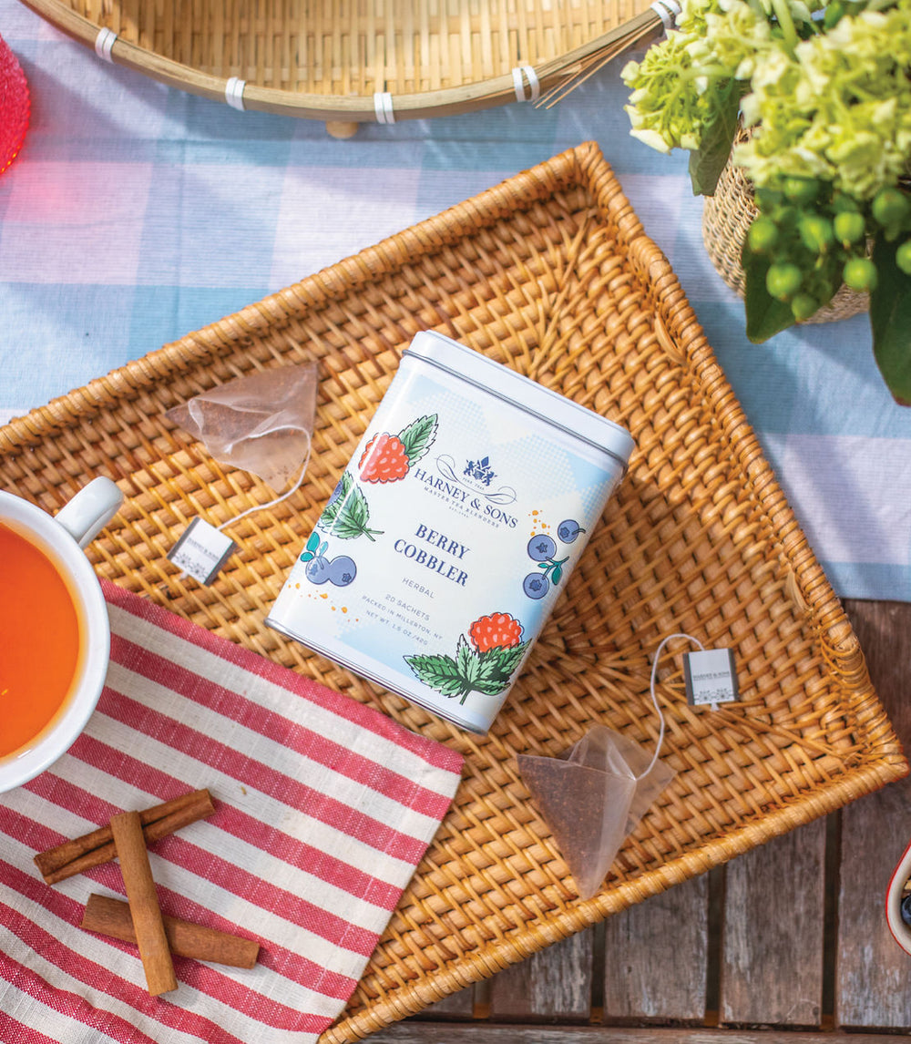 Tea canister labeled Berry Cobbler on a woven tray with teacup, cinnamon sticks, and teabag.