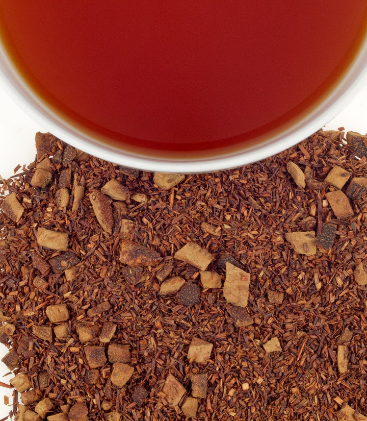 Close-up of dried rooibos tea leaves with a cup of red tea in the background.