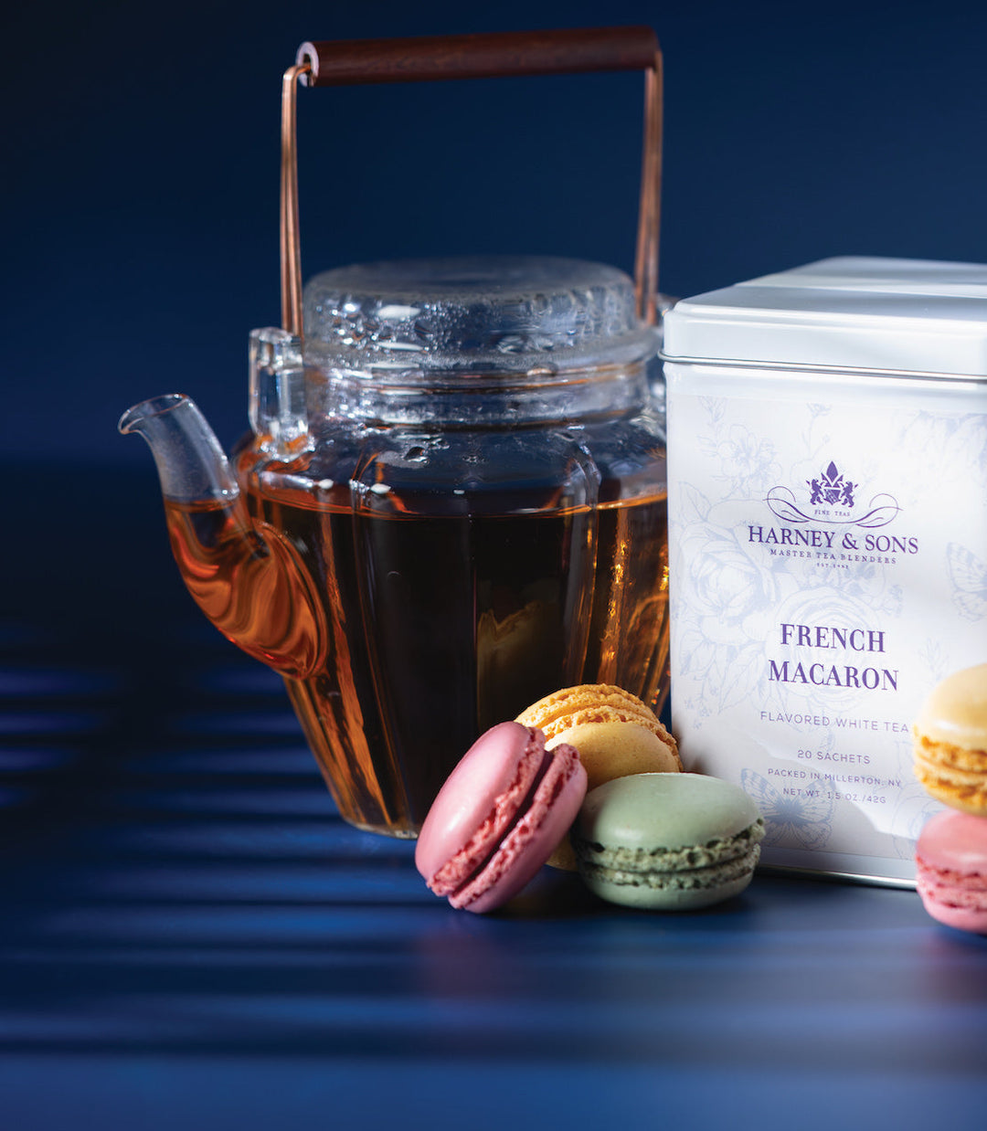 Teapot with tea, colorful macarons, and Harney and Sons French Macaron tea packaging on a dark background.