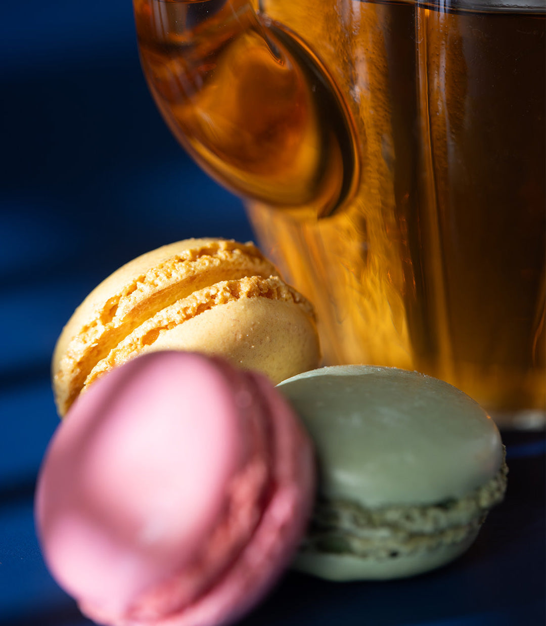 Three macarons in yellow, pink, and green with a glass of amber liquid on a dark background.