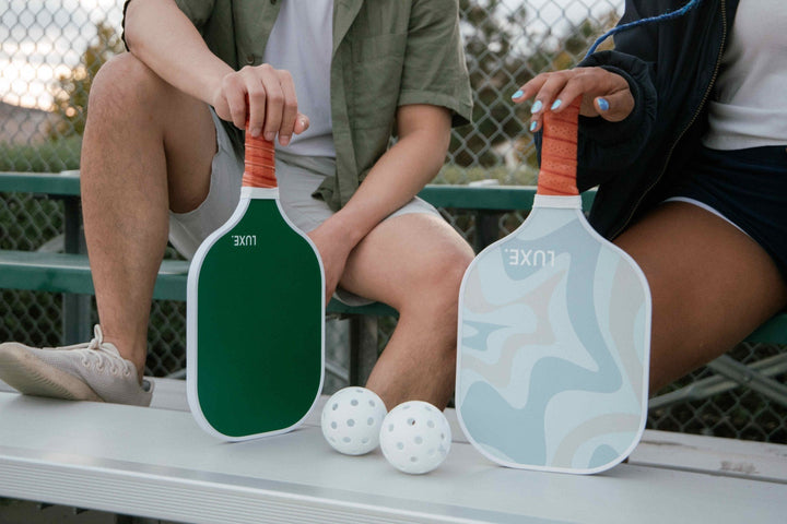 Two people sitting on a bench holding pickleball paddles with balls in front of them.