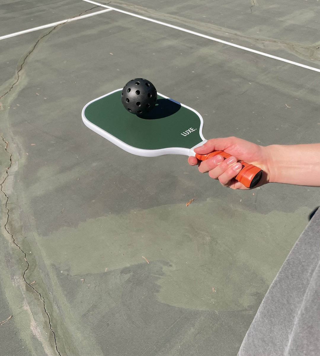 Hand holding a pickleball paddle with a pickleball on a court.