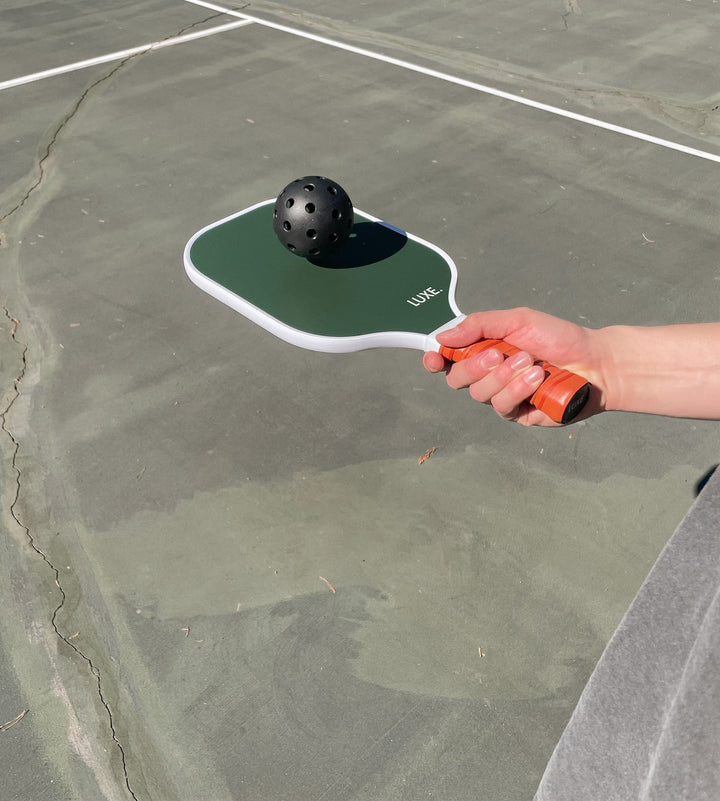Hand holding a pickleball paddle with a pickleball on a court.