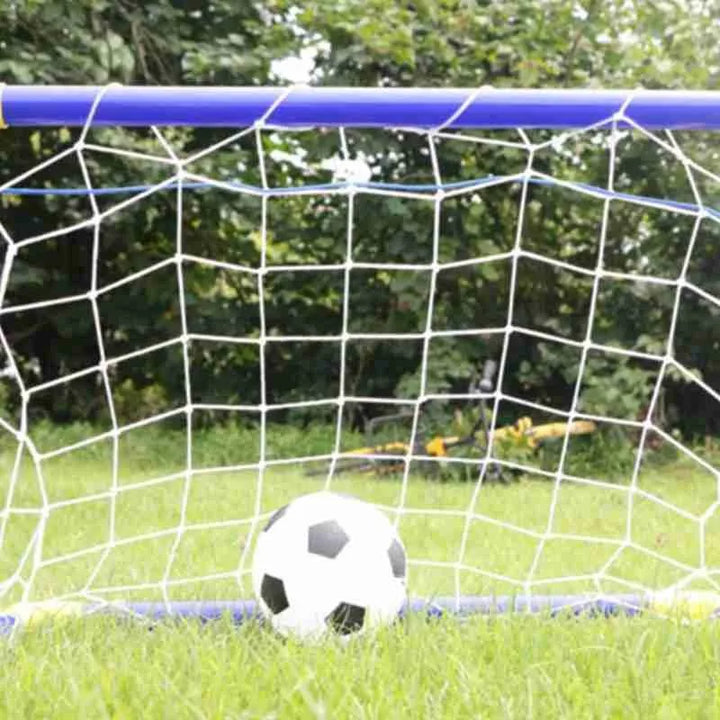 Soccer ball in front of a small soccer goal on grass with trees in the background.