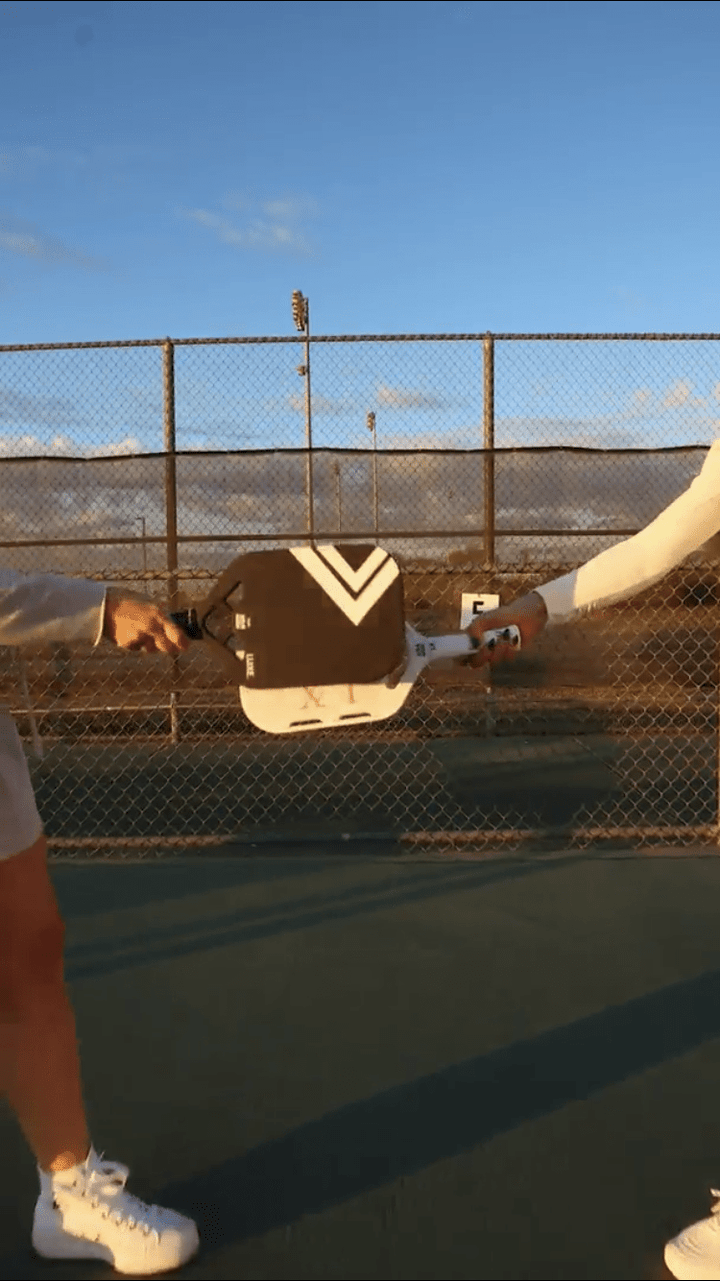 Person holding a tennis racket on a tennis court with a fence and blue sky in the background.
