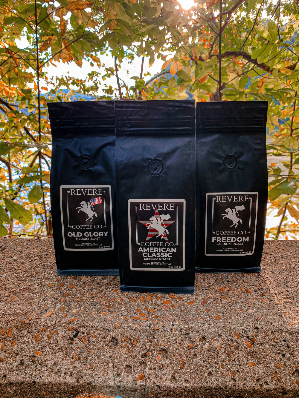 Three black Revere Coffee Co. bags on a stone surface with trees in the background.