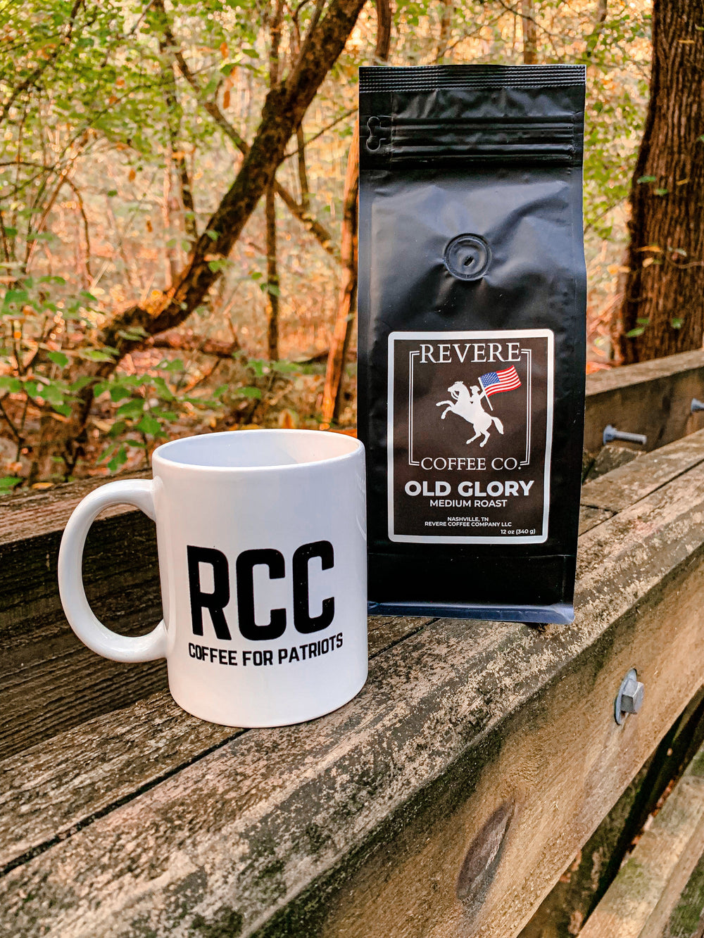 White mug with RCC and black coffee bag labeled Revere Coffee Co. on a wooden surface with a forest background.