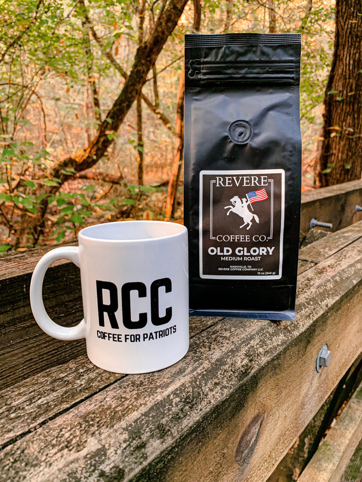 White mug with RCC and black coffee bag labeled Revere Coffee Co. on a wooden surface with a forest background.
