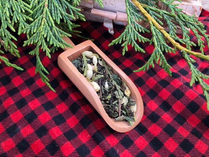 Wooden bowl with dried herbs on a red and black checkered tablecloth with green foliage.