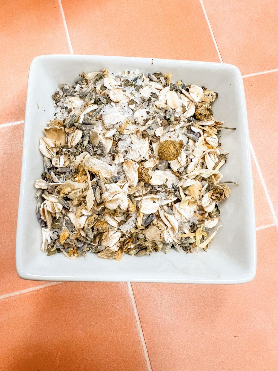 White square bowl filled with dried herbs on a terracotta tiled floor.