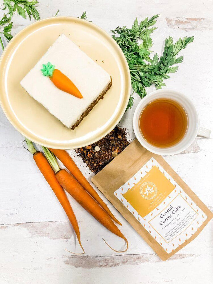 Carrot cake with a decorative carrot on top, surrounded by carrots, a cup of tea, and a package on a white surface.