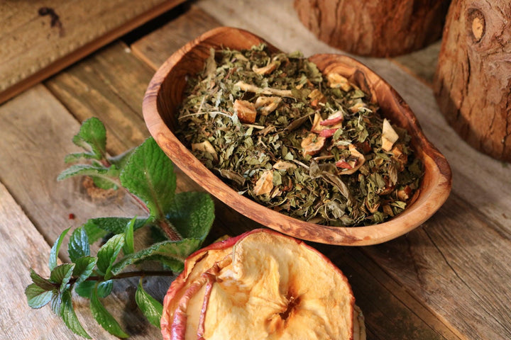 Wooden bowl filled with dried herbs and apples on a wooden surface.