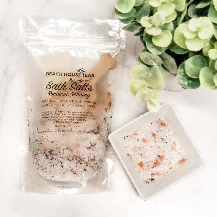 Packaged bath salts with a scoop and small dish of bath salts on a white surface with greenery.