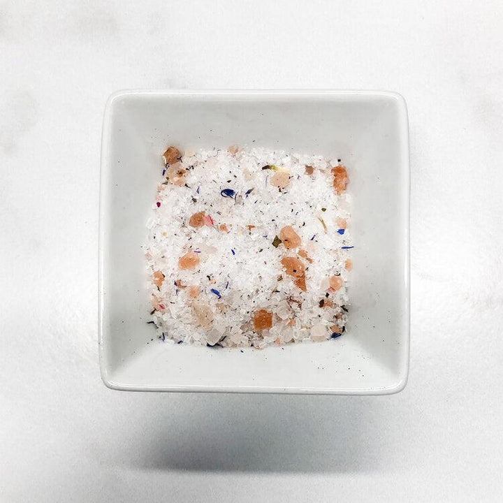 White square bowl filled with multicolored bath salts on a light gray background.