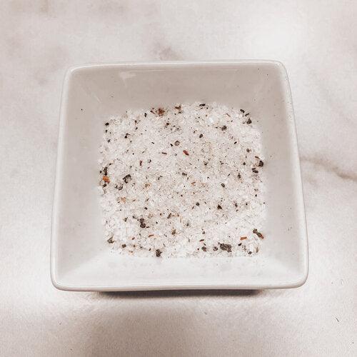 Square white dish with a mixture of white and black bath salts on a light beige background.
