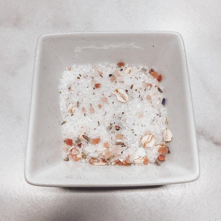 Square white bowl filled with a mixture of white and multicolored bath salts on a light gray background.