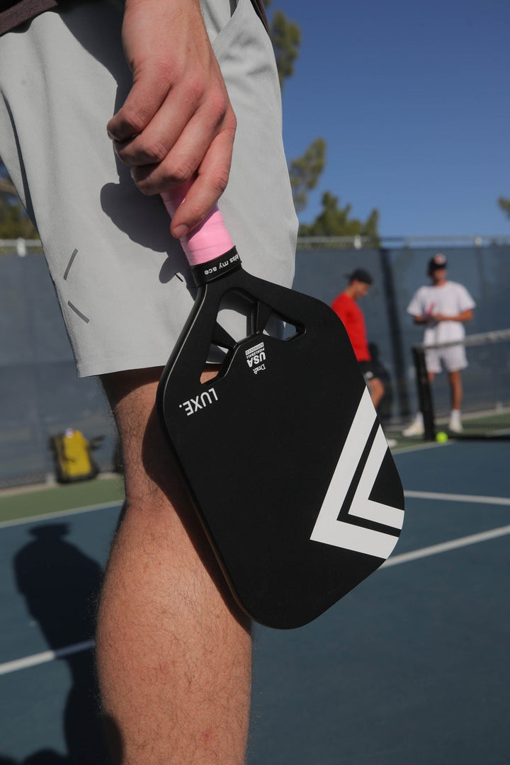 Person holding a black and pink pickleball paddle on a tennis court with other players in the background.