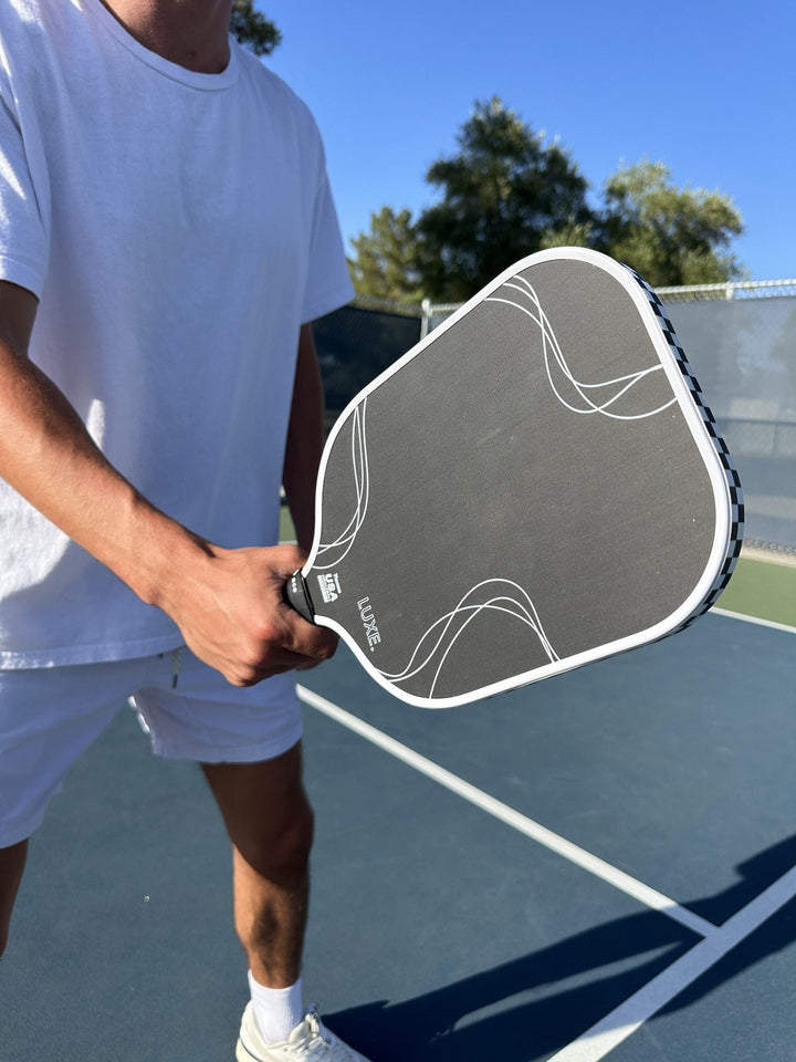Person holding a pickleball paddle on a court with trees in the background.