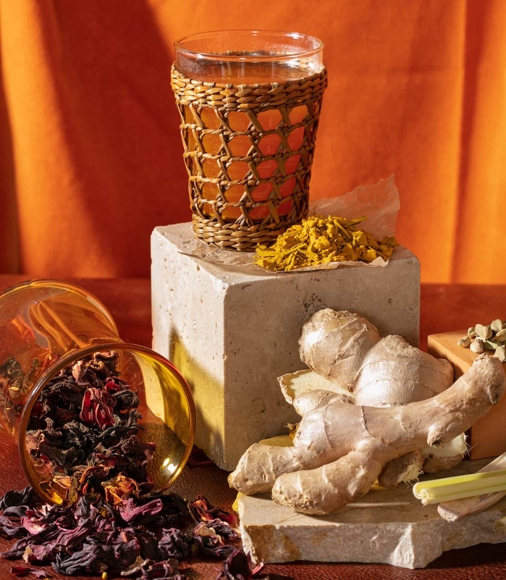 Glass with a woven sleeve on a stone block, surrounded by dried herbs and ginger roots against an orange background.