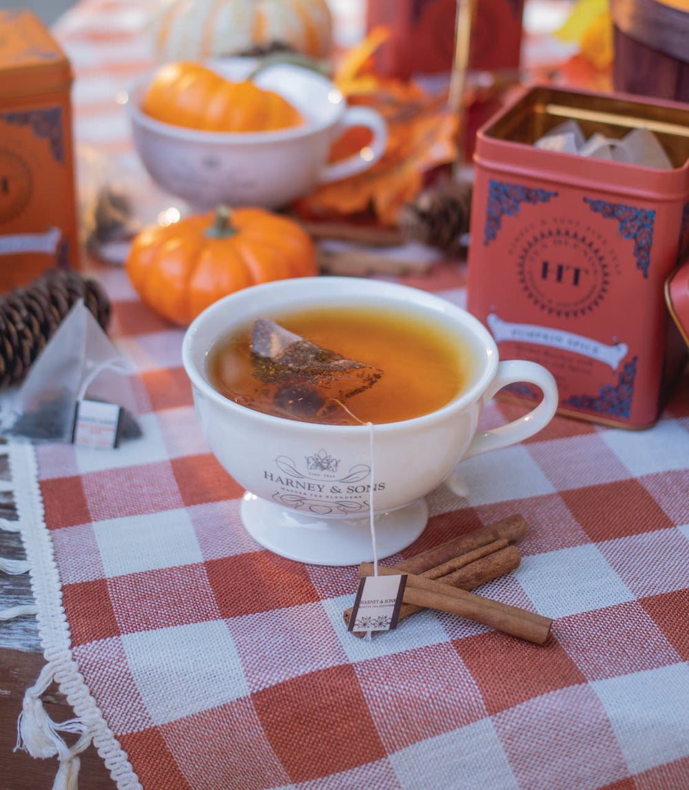 Teacup with Harney and Sons tea bag on a checkered tablecloth with pumpkins and tea boxes in the background.
