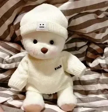White teddy bear wearing a knitted sweater and hat on a striped fabric background.