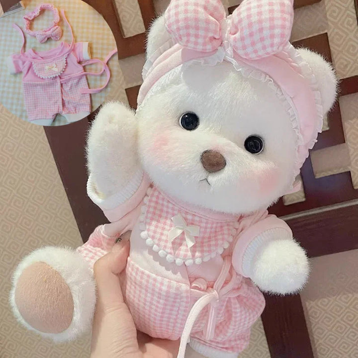 White teddy bear with pink checkered outfit and bow held by a hand, with baby clothes in the background.