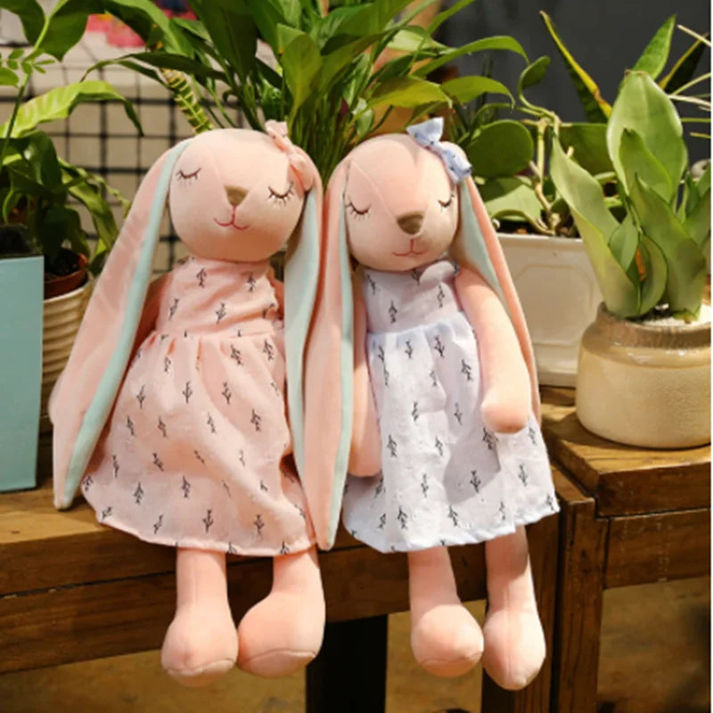 Two plush bunny toys in dresses sitting on a wooden surface with plants in the background.