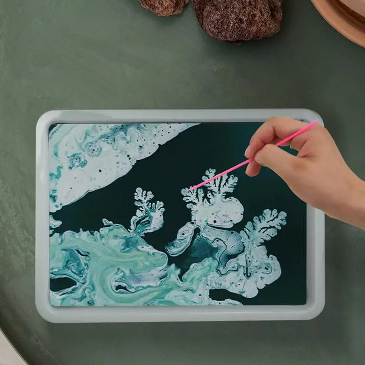 Hand holding a pink straw over a tablet displaying an underwater scene with marine life on a green surface.