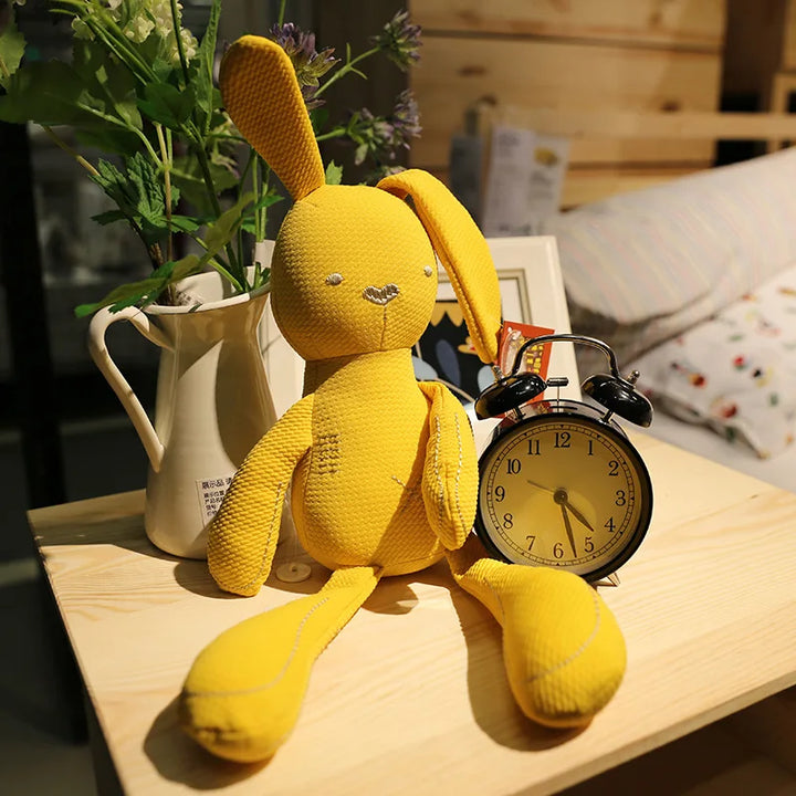 Yellow plush bunny toy next to a vintage alarm clock on a wooden surface with plants in the background.