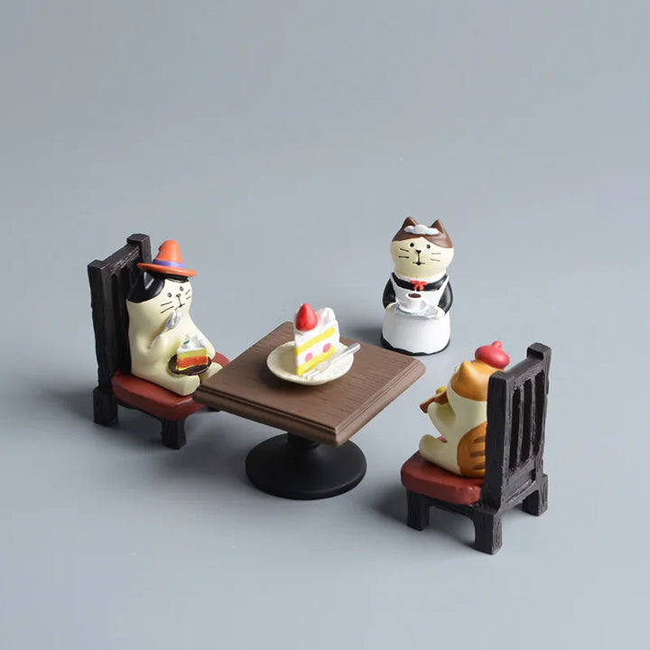 Three cat-shaped figurines around a table with a cake on a gray background.