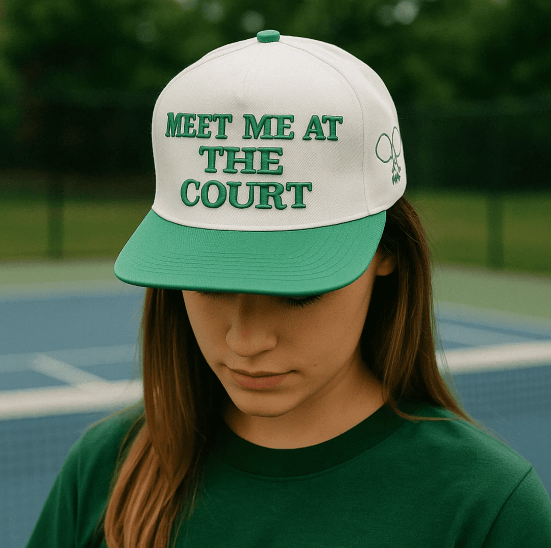 Person wearing a cap with Meet Me at the Court text on a tennis court.