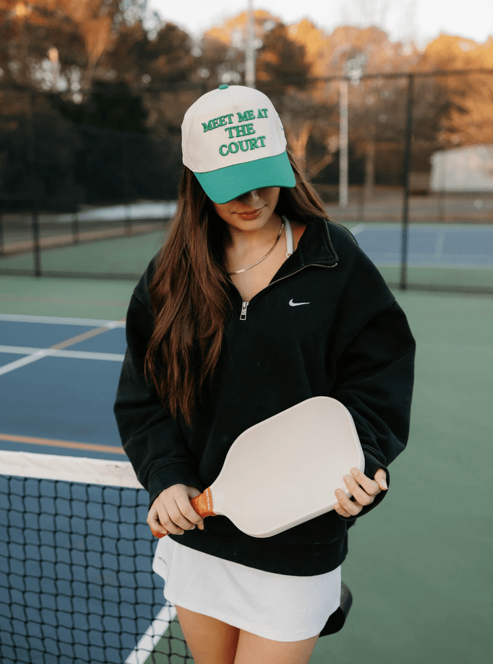 Woman on a tennis court wearing a cap and holding a paddle.