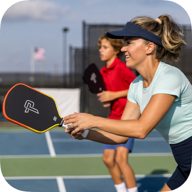 Woman holding a pickleball paddle on a tennis court with another player in the background.