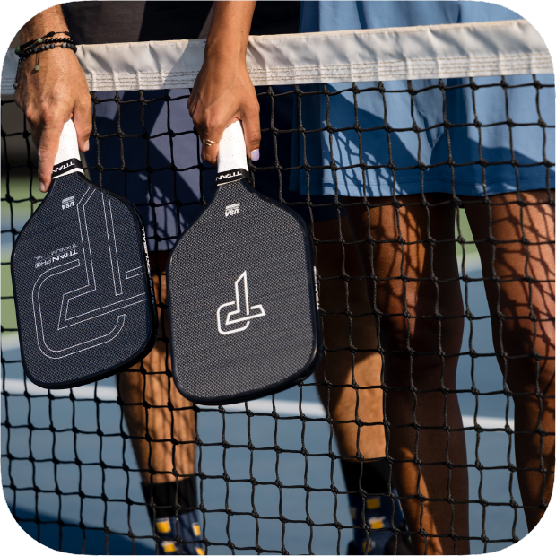 Person holding two pickleball paddles behind a net on a court.